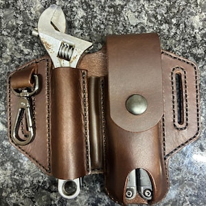 May include: A brown leather belt holster featuring a silver metal swivel clip on the left, a central sleeve holding a silver adjustable wrench, and a right-side pouch with a snap button closure containing a multi-tool with visible silver screws.
