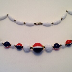 May include: A white beaded necklace with red and navy blue accents. The necklace features a variety of bead shapes, including elongated white beads and round beads in red, white, and navy blue. The necklace is secured with a magnetic clasp.