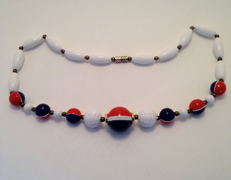 May include: A white beaded necklace with red and navy blue accents. The necklace features a variety of bead shapes, including elongated white beads and round beads in red, white, and navy blue. The necklace is secured with a magnetic clasp.