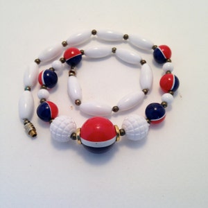 May include: A vintage necklace with white, red, and navy blue beads. The necklace features a mix of bead shapes, including elongated ovals, round, and textured spherical beads. Gold-tone metal accents are used throughout the design.
