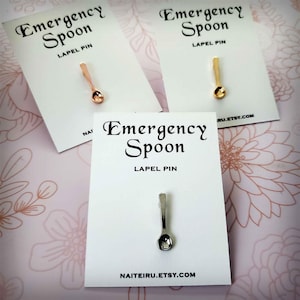 May include: Three cards with the text "Emergency Spoon" and "Lapel Pin" printed on them. Each card features a small spoon-shaped pin in a different color: gold, rose gold, and silver.