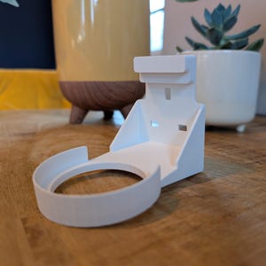 May include: A white, 3D-printed wall mount for a smart home device. The mount has a circular base and a rectangular top section. The device holder is on a wooden surface, with a yellow object and a white pot with a plant in the background.