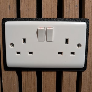 May include: A white double electrical socket with two switches marked "ON". The socket is set in a black frame and mounted on a wooden panel.
