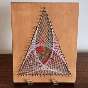May include: A rectangular wooden board featuring a string art design in the shape of a triangle. The design is created with white, red, and green threads wrapped around small metal nails, forming intricate geometric patterns within the triangle.