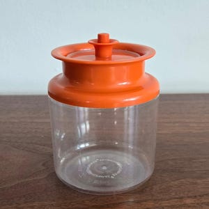 May include: A clear, cylindrical storage canister featuring a bright orange lid with a small, central knob handle. The container has a wide, flared rim design and a transparent body, allowing for a clear view of the interior space within the jar.