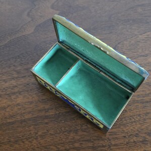 Puede incluir: Una caja de baratijas rectangular abierta con tapa con bisagras. La caja es de color dorado con detalles de esmalte azul y amarillo. El interior est&aacute; forrado con tela verde y dividido en dos compartimentos. La caja est&aacute; sobre una superficie de madera.