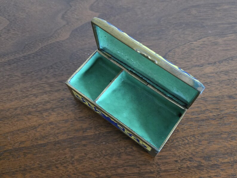 Puede incluir: Una caja de baratijas rectangular abierta con tapa con bisagras. La caja es de color dorado con detalles de esmalte azul y amarillo. El interior est&aacute; forrado con tela verde y dividido en dos compartimentos. La caja est&aacute; sobre una superficie de madera.