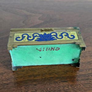Puede incluir: Una peque&ntilde;a caja rectangular con tapa abisagrada. La caja tiene un marco de lat&oacute;n y est&aacute; decorada con dise&ntilde;os de esmalte azul y amarillo. La parte delantera de la caja es verde con la palabra "CHINA" en letras rojas. Es probable que sea un art&iacute;culo antiguo o vintage.