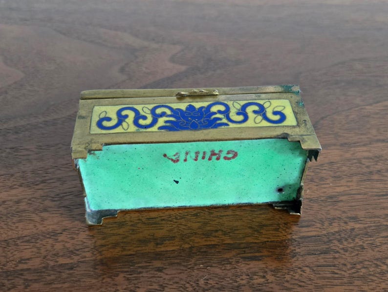 Puede incluir: Una peque&ntilde;a caja rectangular con tapa abisagrada. La caja tiene un marco de lat&oacute;n y est&aacute; decorada con dise&ntilde;os de esmalte azul y amarillo. La parte delantera de la caja es verde con la palabra "CHINA" en letras rojas. Es probable que sea un art&iacute;culo antiguo o vintage.