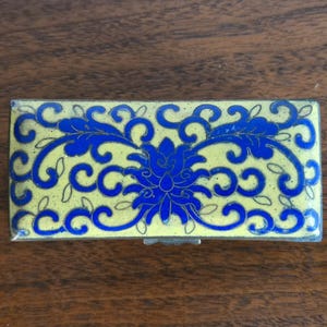 Puede incluir: Caja rectangular para baratijas con fondo amarillo y dise&ntilde;o floral azul. La caja tiene un marco de metal plateado y una tapa con bisagras. El dise&ntilde;o floral presenta una flor central y patrones arremolinados.