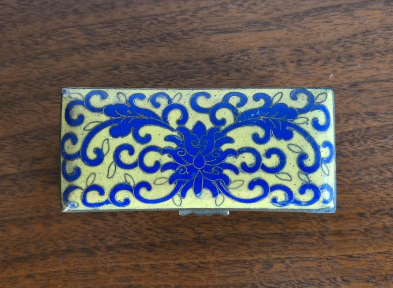 Puede incluir: Caja rectangular para baratijas con fondo amarillo y dise&ntilde;o floral azul. La caja tiene un marco de metal plateado y una tapa con bisagras. El dise&ntilde;o floral presenta una flor central y patrones arremolinados.