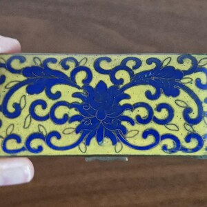 Puede incluir: Caja rectangular para baratijas con fondo amarillo y dise&ntilde;o floral azul. La caja tiene tapa con bisagras y cierre met&aacute;lico. El dise&ntilde;o floral presenta una flor central y patrones arremolinados.