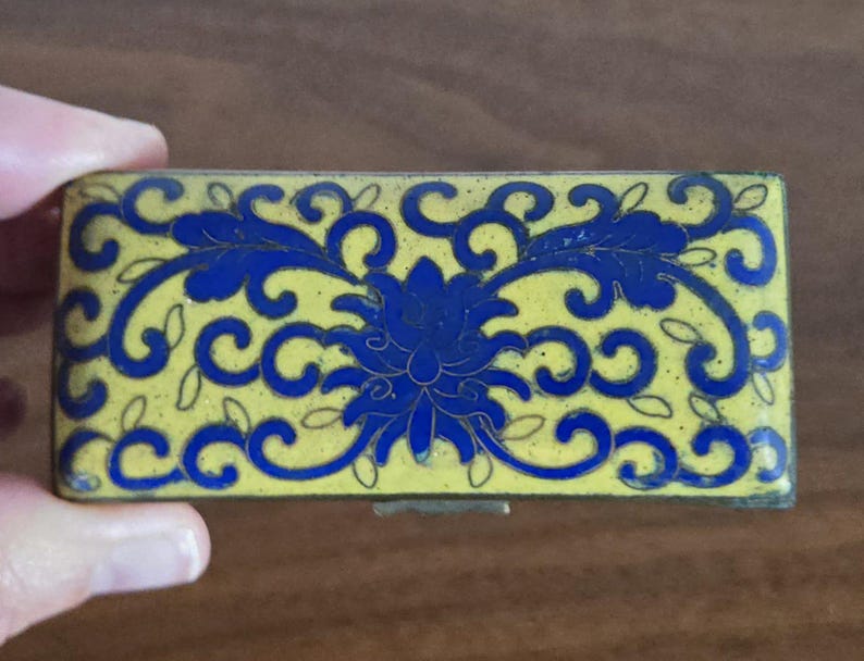 Puede incluir: Caja rectangular para baratijas con fondo amarillo y dise&ntilde;o floral azul. La caja tiene tapa con bisagras y cierre met&aacute;lico. El dise&ntilde;o floral presenta una flor central y patrones arremolinados.