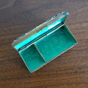 Puede incluir: Una caja rectangular antigua abierta con tapa abisagrada. La caja tiene un marco de metal dorado con un interior turquesa y una base verde vibrante. La tapa est&aacute; decorada con detalles azules y amarillos. La caja est&aacute; sobre una superficie de madera.