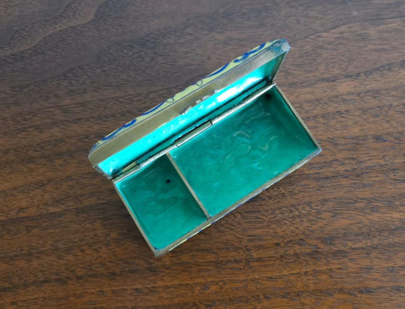 Puede incluir: Una caja rectangular antigua abierta con tapa abisagrada. La caja tiene un marco de metal dorado con un interior turquesa y una base verde vibrante. La tapa est&aacute; decorada con detalles azules y amarillos. La caja est&aacute; sobre una superficie de madera.