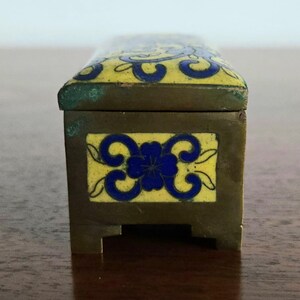 Puede incluir: Peque&ntilde;a caja rectangular de lat&oacute;n con tapa con bisagras. La tapa y el panel frontal presentan dise&ntilde;os florales azules sobre fondo amarillo. Probablemente para guardar objetos peque&ntilde;os, medidas en cent&iacute;metros.