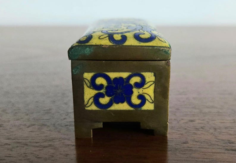 Puede incluir: Peque&ntilde;a caja rectangular de lat&oacute;n con tapa con bisagras. La tapa y el panel frontal presentan dise&ntilde;os florales azules sobre fondo amarillo. Probablemente para guardar objetos peque&ntilde;os, medidas en cent&iacute;metros.