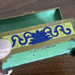 Puede incluir: Una caja de baratijas rectangular antigua y abierta con tapa con bisagras. El interior de la caja es verde y tiene un dise&ntilde;o floral amarillo y azul en la parte delantera. La palabra "CHINA" es visible en la parte inferior.