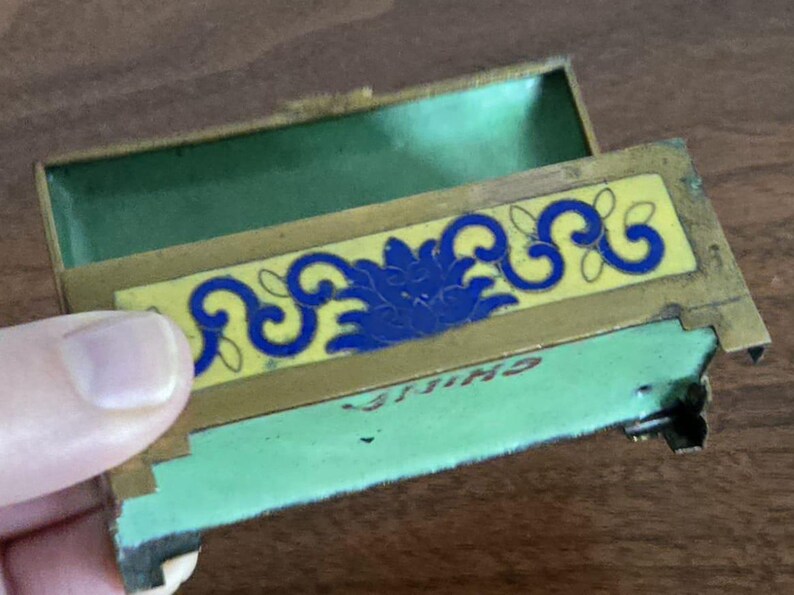Puede incluir: Una caja de baratijas rectangular antigua y abierta con tapa con bisagras. El interior de la caja es verde y tiene un dise&ntilde;o floral amarillo y azul en la parte delantera. La palabra "CHINA" es visible en la parte inferior.