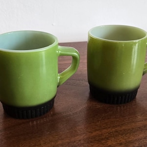 May include: Two matching mugs with a smooth, light green upper body and a dark, ribbed base. Each mug features a sturdy, integrated handle on the side and a wide, open top. The interior of the mugs is a pale, off-white color, contrasting with the vibrant green exterior finish.