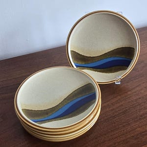 May include: A stack of beige ceramic plates with a brown and blue wave design. The plates have a gold rim and are stacked on a wooden surface. The plates are part of a dinnerware set.