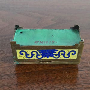 Puede incluir: Peque&ntilde;a caja rectangular antigua con tapa y laterales verdes. La caja tiene una banda amarilla con dise&ntilde;os florales azules. La palabra "CHINA" est&aacute; impresa en la parte superior. La caja es de metal y est&aacute; sobre una superficie de madera.