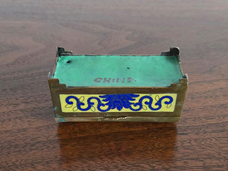 Puede incluir: Peque&ntilde;a caja rectangular antigua con tapa y laterales verdes. La caja tiene una banda amarilla con dise&ntilde;os florales azules. La palabra "CHINA" est&aacute; impresa en la parte superior. La caja es de metal y est&aacute; sobre una superficie de madera.