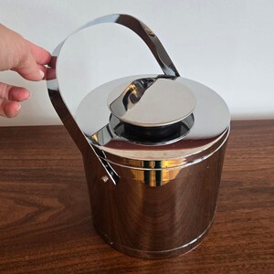 May include: A cylindrical ice bucket with a polished chrome finish, featuring a matching lid with a central knob and a tall, arched swing handle attached to the sides. The reflective metal surface shows a smooth, seamless exterior design.
