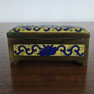 Puede incluir: Peque&ntilde;a caja rectangular para baratijas con marco de lat&oacute;n y tapa y laterales de esmalte amarillo. El esmalte est&aacute; decorado con motivos florales azules. La caja mide aproximadamente 7.6 cm de largo.