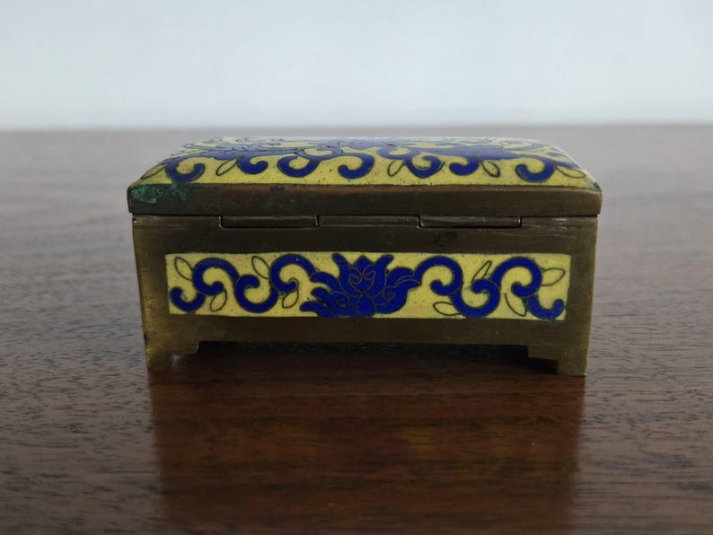 Puede incluir: Peque&ntilde;a caja rectangular para baratijas con marco de lat&oacute;n y tapa y laterales de esmalte amarillo. El esmalte est&aacute; decorado con motivos florales azules. La caja mide aproximadamente 7.6 cm de largo.