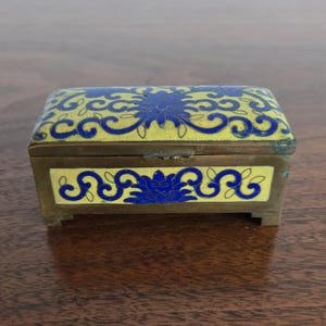Puede incluir: Una peque&ntilde;a caja rectangular de lat&oacute;n con tapa con bisagras. La caja presenta dise&ntilde;os florales azules sobre un fondo de esmalte amarillo. La caja est&aacute; sobre una superficie de madera.