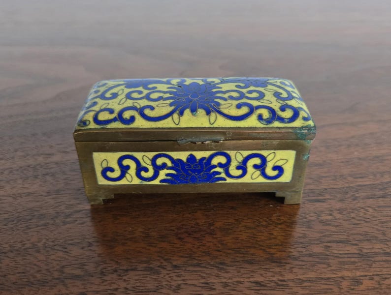 Puede incluir: Una peque&ntilde;a caja rectangular de lat&oacute;n con tapa con bisagras. La caja presenta dise&ntilde;os florales azules sobre un fondo de esmalte amarillo. La caja est&aacute; sobre una superficie de madera.