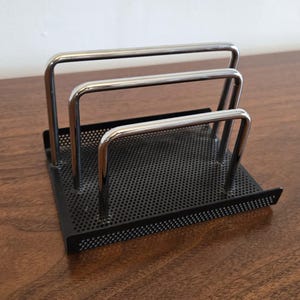 May include: A black metal desk organizer with three chrome-colored bars. The organizer has a perforated base and is designed to hold documents or other office supplies. The item is on a wooden surface.