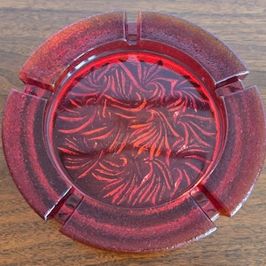 May include: A circular red glass ashtray featuring a textured, frosted outer rim with three evenly spaced notches. The center basin displays a deep red, swirling, fan-like pattern embossed into the glass, creating a distinct visual contrast.