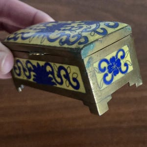 Puede incluir: Peque&ntilde;a caja rectangular para baratijas con tapa con bisagras. La caja es de color dorado con esmalte amarillo y dise&ntilde;os florales azules. Tiene peque&ntilde;os pies y est&aacute; sujeta por una mano.