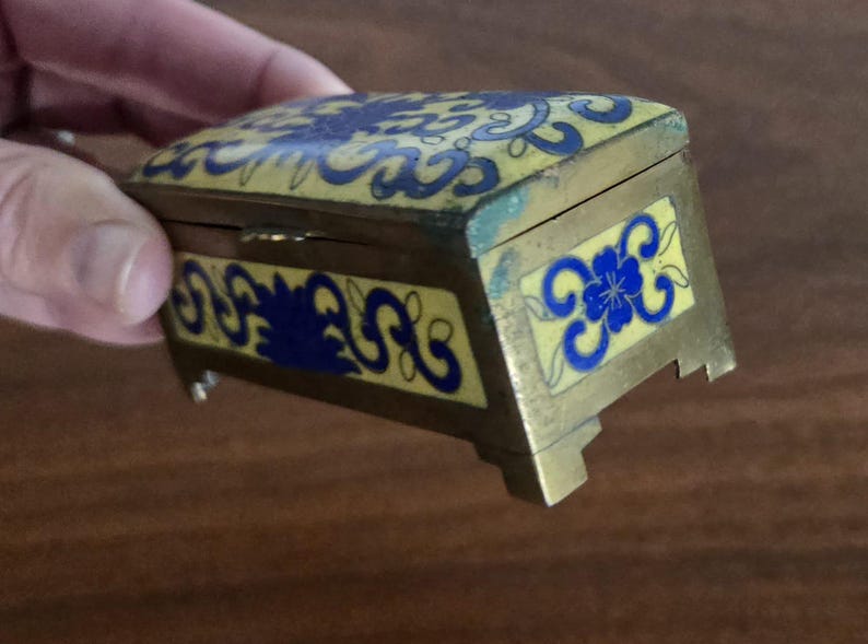 Puede incluir: Peque&ntilde;a caja rectangular para baratijas con tapa con bisagras. La caja es de color dorado con esmalte amarillo y dise&ntilde;os florales azules. Tiene peque&ntilde;os pies y est&aacute; sujeta por una mano.
