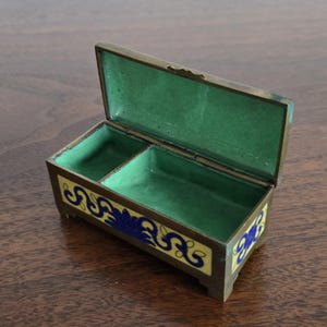 Puede incluir: Caja de joyer&iacute;a rectangular abierta, con tapa con bisagras. El interior es verde y el exterior est&aacute; decorado con patrones florales amarillos y azules. La caja es de metal y tiene peque&ntilde;os pies.