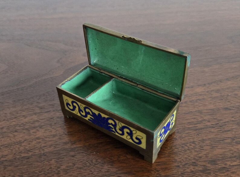 Puede incluir: Caja de joyer&iacute;a rectangular abierta, con tapa con bisagras. El interior es verde y el exterior est&aacute; decorado con patrones florales amarillos y azules. La caja es de metal y tiene peque&ntilde;os pies.