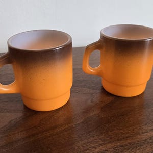 May include: Two stackable mugs featuring a gradient design that transitions from a dark brown rim to a bright orange base. Each mug has a D-shaped handle and a smooth, glossy finish, showcasing a distinct color fade from top to bottom.