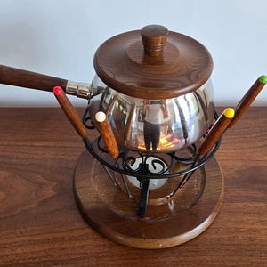 May include: A stainless steel fondue pot with a dark wood lid and handle, resting on a black metal stand with a circular wood base. The set includes several fondue forks with wood handles featuring red, white, yellow, and green colored tips.