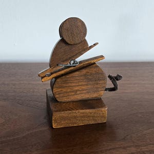 May include: A wooden mouse-shaped clothespin holder. The mouse is made of dark brown wood with a clothespin for the mouth and a pipe cleaner tail. The holder sits on a small wooden base.
