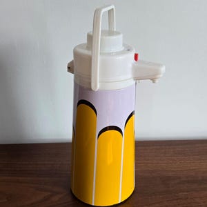 May include: A vintage white and yellow insulated beverage dispenser with a handle and a spout. The body of the dispenser is decorated with a yellow and purple pattern. The dispenser is designed to keep beverages hot or cold.