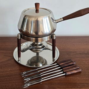 May include: A stainless steel fondue pot with a lid, featuring a long wooden handle and a matching wooden knob. The pot rests on a metal stand with wooden legs, accompanied by six metal fondue forks with dark wooden handles arranged in a row.