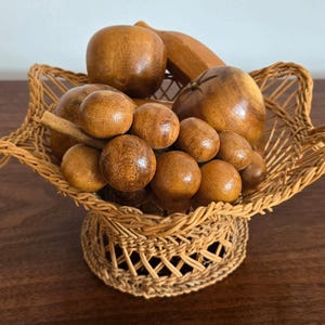 May include: A woven wicker basket with a scalloped rim containing a collection of carved wooden fruit, including a smooth apple, a textured bunch of grapes, and a banana, all featuring natural brown wood grain tones and polished finishes.