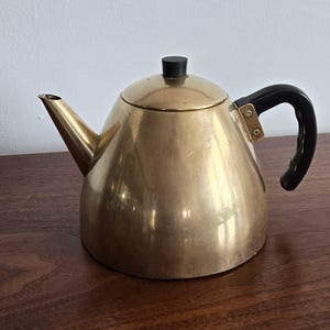 May include: A brass teapot with a black handle and lid. The teapot has a rounded body and a long spout. The handle is curved and attached to the side of the teapot. The lid is small and round. The teapot is sitting on a wooden surface.