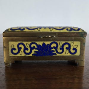 Puede incluir: Peque&ntilde;a caja rectangular de lat&oacute;n con tapa abisagrada. Decorada con esmalte amarillo y dise&ntilde;os florales azules. Descansa sobre cuatro peque&ntilde;os pies. Ideal para guardar joyas u objetos peque&ntilde;os. Mide unos 7 cm de largo.