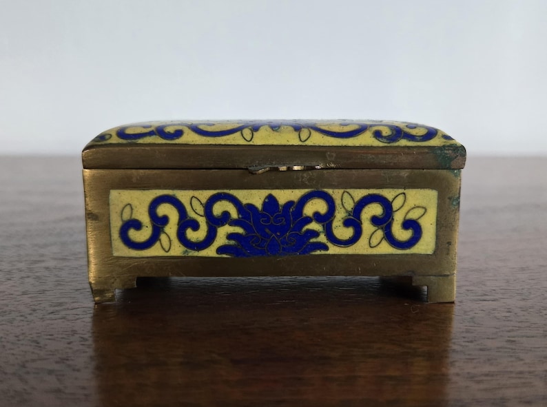 Puede incluir: Peque&ntilde;a caja rectangular de lat&oacute;n con tapa abisagrada. Decorada con esmalte amarillo y dise&ntilde;os florales azules. Descansa sobre cuatro peque&ntilde;os pies. Ideal para guardar joyas u objetos peque&ntilde;os. Mide unos 7 cm de largo.