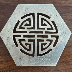 May include: A hexagonal brass trivet featuring a central cut-out geometric Shou symbol. The metal surface is decorated with engraved floral patterns and a fine stippled texture around the edges, showcasing a metallic gold-toned finish.