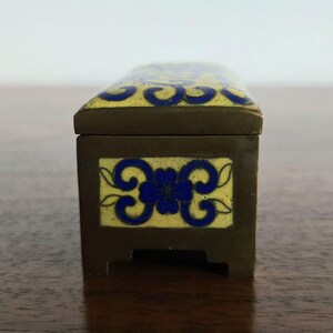 Puede incluir: Una caja rectangular con tapa con bisagras, de color marr&oacute;n oscuro y adornada con dise&ntilde;os florales esmaltados amarillos y azules. La caja est&aacute; sobre una superficie de madera. El dise&ntilde;o de la caja recuerda a los estilos antiguos.