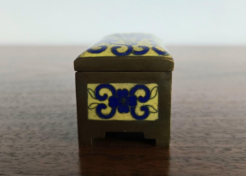 Puede incluir: Una caja rectangular con tapa con bisagras, de color marr&oacute;n oscuro y adornada con dise&ntilde;os florales esmaltados amarillos y azules. La caja est&aacute; sobre una superficie de madera. El dise&ntilde;o de la caja recuerda a los estilos antiguos.